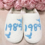 Fluffy white slippers with blue 1989 text and bird graphics, set against pink flowers.