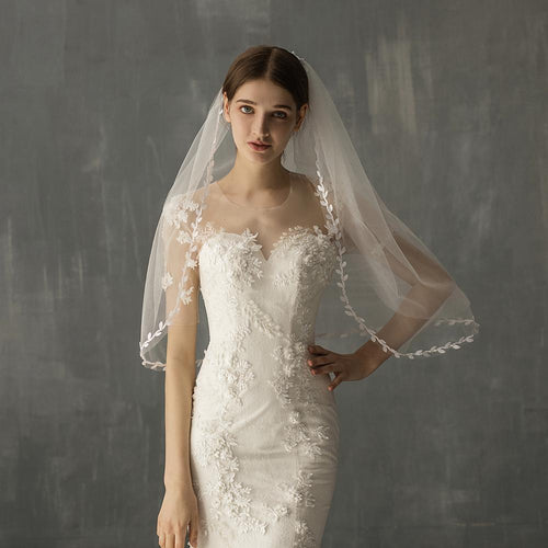 A woman wearing a white lace wedding dress and a sheer tulle veil with leaf embroidery along the edge.