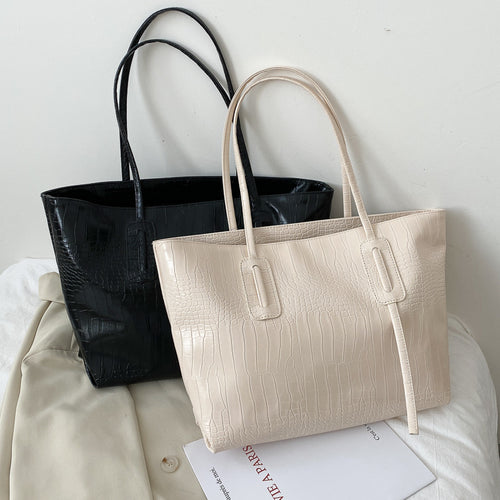 Two croc-embossed leather tote bags in black and cream, with shoulder straps, arranged on a bed.