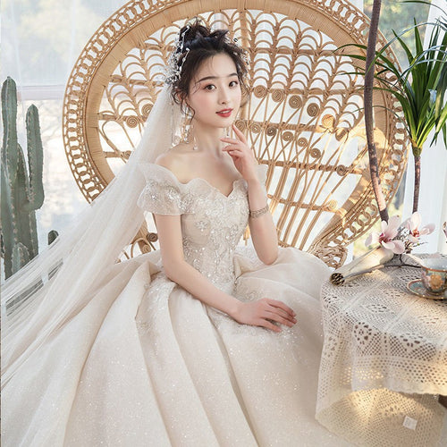 A bride in an off-the-shoulder sparkling ivory ballgown seated in a wicker chair with a matching veil.