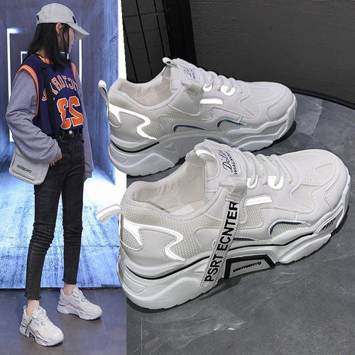 White chunky platform sneakers with reflective panels, lace-up front, and branded strap, shown paired with a casual outfit.