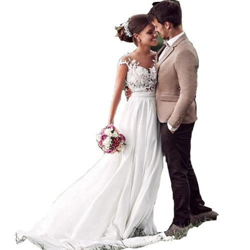 Bride in a white chiffon gown with a lace bodice and groom in a beige suit facing each other holding flowers.