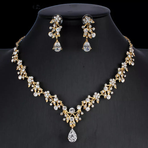 Gold-colored necklace and earring set with floral motifs and clear zirconia crystals on a black stand.