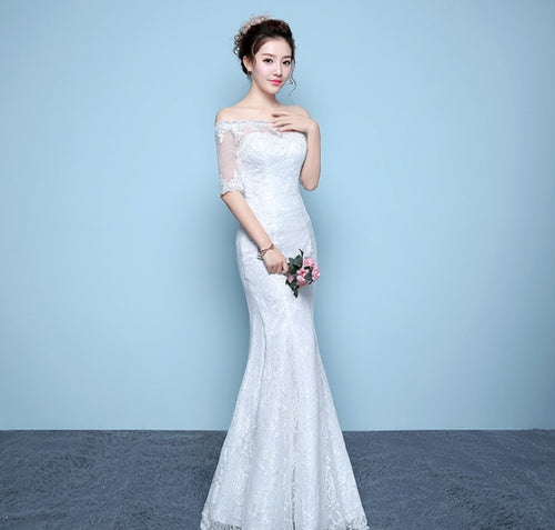 A woman posing in a white off-shoulder lace mermaid wedding dress holding pink flowers.