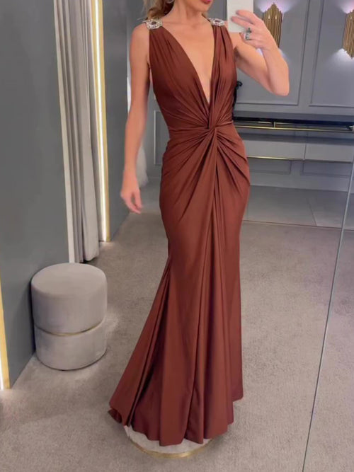 A woman wearing a draped, floor-length brown silk evening gown with a plunging neckline and embellished straps.