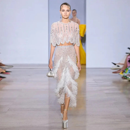 A model on a runway wears a two-piece, beaded outfit adorned with white feathers and sheer fabric.