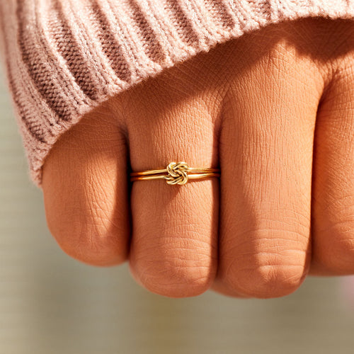 A gold double knot ring worn on a finger, near the cuff of a pink knitted sweater.