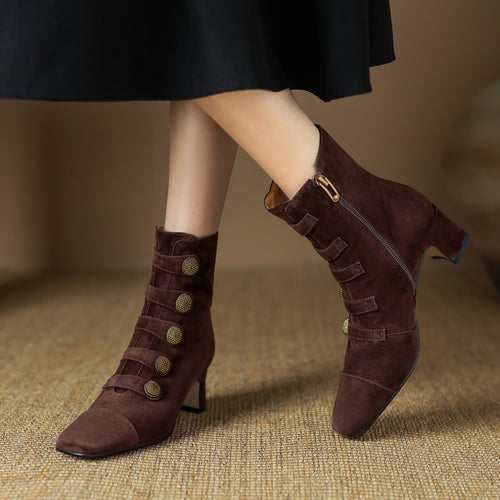 Brown suede ankle boots with block heels, decorative straps, and round textured brass buttons.