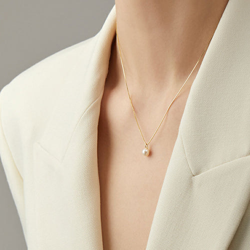 A gold chain necklace with a single round white pearl pendant worn on a person in a white blazer.