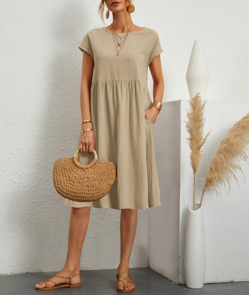 A model wears a beige, short-sleeved round-neck dress with a gathered waist, holding a straw bag.