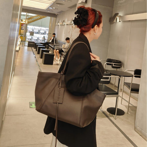 A person wearing a brown leather shoulder tote bag with thin straps and a hanging luggage tag.