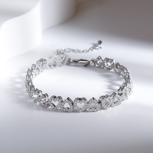 Silver tennis bracelet featuring linked heart-shaped charms paved with sparkling stones on a white surface.