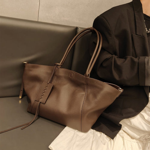 Brown leather shoulder tote bag with double straps and a hanging luggage tag on a sofa.