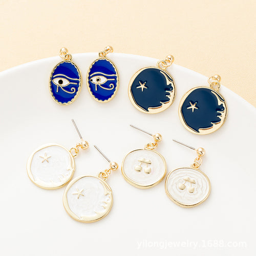 Four pairs of gold-rimmed drop earrings featuring blue and white enamel designs of evil eyes, moons, and cherries on a white plate.