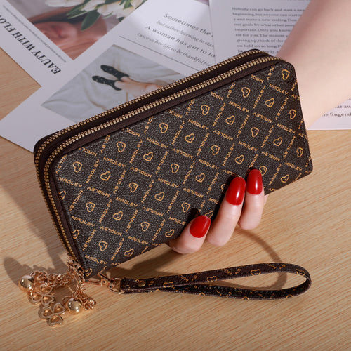 Brown leather double-zipper wallet with gold monogram print, wrist strap, and gold charm held by a hand.