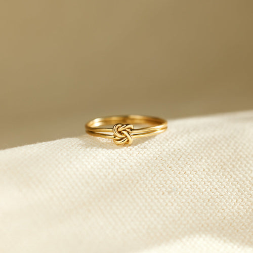 A gold-colored metal ring with a double band tied into a central knot, resting on a textured fabric surface.