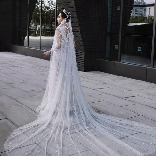 A long white tulle wedding veil adorned with scattered pearls, worn by a bride outdoors.