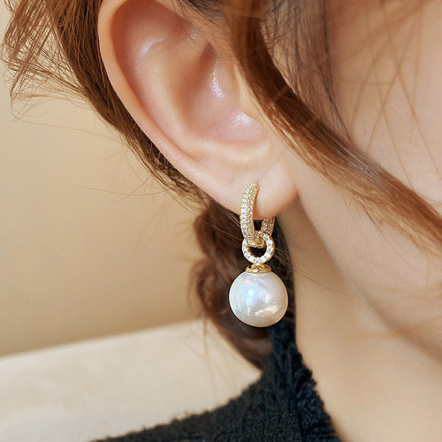 Gold hoop earring encrusted with crystals, featuring a hanging white pearl drop on an ear.